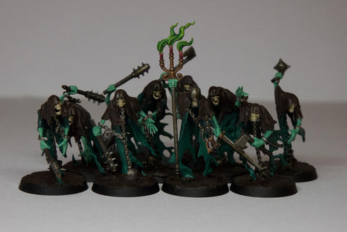 Ten blue green ghosts with dark brown capes, with various weapons. Their bodies are a dark blue green while their arms are a ghoulish green, their heads are pale bone. Most have various rusted and damaged weapons, the one in the centre is holding a candle stand with pink candles and pale green flames. They are all stood on muddy bases  