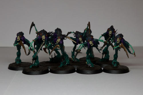 Ten hunched over floating blue green ghosts with scythes for hands, bleached skulls and black hair with flowers in it. They have purple cloaks and the flowers in their hair a mix of purple, red and blue. Their scythes are made from different metals, with the leader having two pairs of arms. They are floating over a muddy base