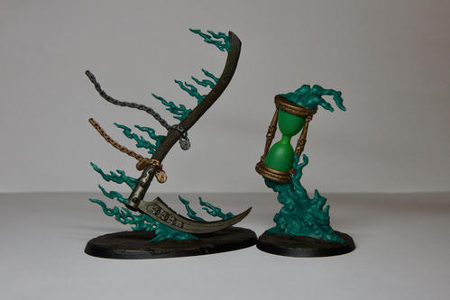 Two Warhammer spell models. The left is a large wooden and metal scythe trailing metal chains and green spirits from its handle. The right is a large time green and gold hourglass being held up by a swam of green smoke with skulls in it. They are both on drak brown muddy basses 