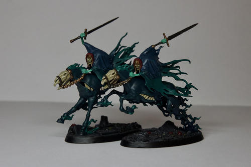 A pair of Warhammer Nighthaunt cavalry models. Both steeds are skeletal with dark blue-green skin, their skulls are exposed and in places their rib cages are showing. Riding them are two blue-green ghosts with dark blue capes and green skin. Both have brass helms and are holding their swords above and behind them. They are on dark stoney bases