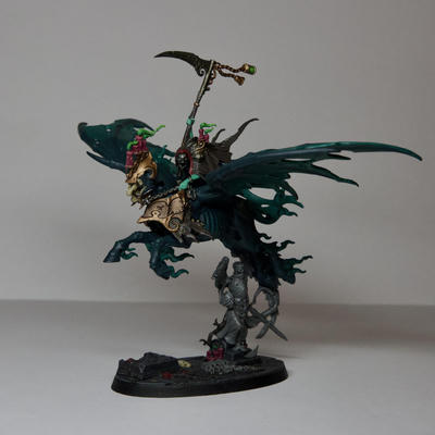 A large flying cavalry Warhammer model. Reikenor is a blue-green ghost with a brown cloak riding on a winged horse. He is holding up a large steel scythe with brass trinkets flying off the back. The horse is a dark blue-green with lighter green wisps of smoke coming off of him. It also has large wings which are also blue-green, and is wearing some brass and gold plate on its front and head. Its skull is visible below the helm, and the helm has various dark pink candles on it with green smoke. He is flying above a stone statue of a soldier on top of a dark stone base