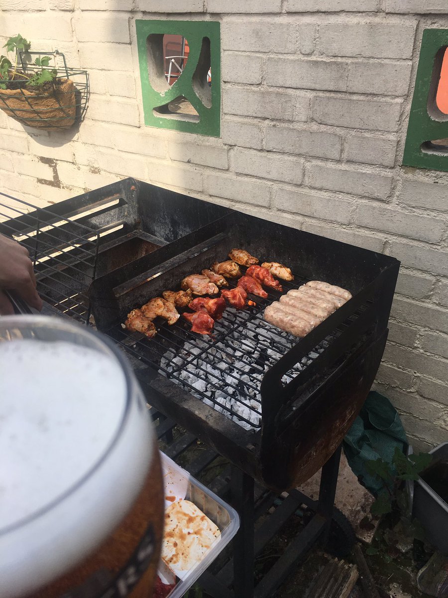 Photo of bbq with various flavoured chicken wings and some sausages. In the background is a white wall. In the lower right is a glass with a frothy cider in it
